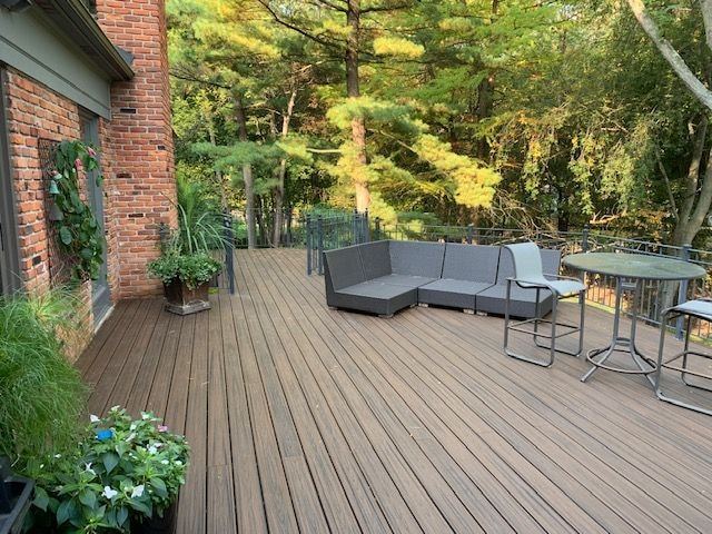 Composite deck with a gray sectional and bistro table beside a brick chimney, woods behind